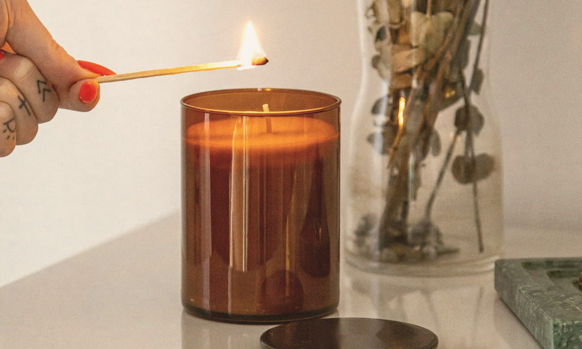 Une main allume une bougie dans un verre ambré avec une allumette